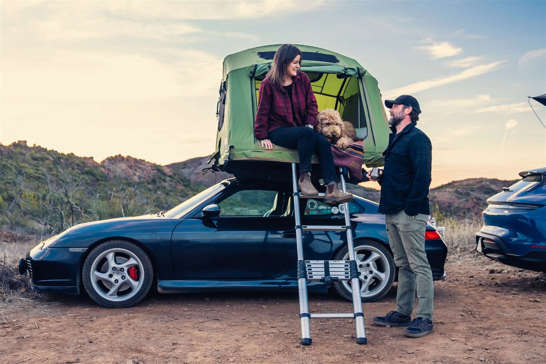 Brock Keen talking to his wife Sara, who sits in a roof tent with their dog Lucy on top of their Porsche 996 Carrera 4S